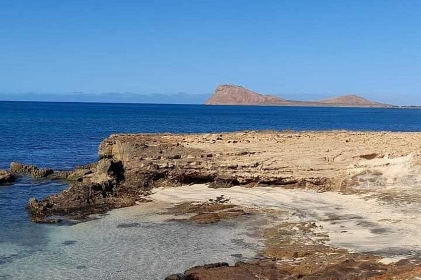 A coastal landscape with rocky terrain, clear blue sea, and distant mountains. 4