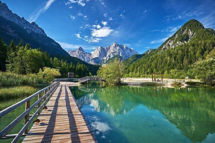 A wooden bridge over a clear lake, surrounded by mountains and greenery. 1
