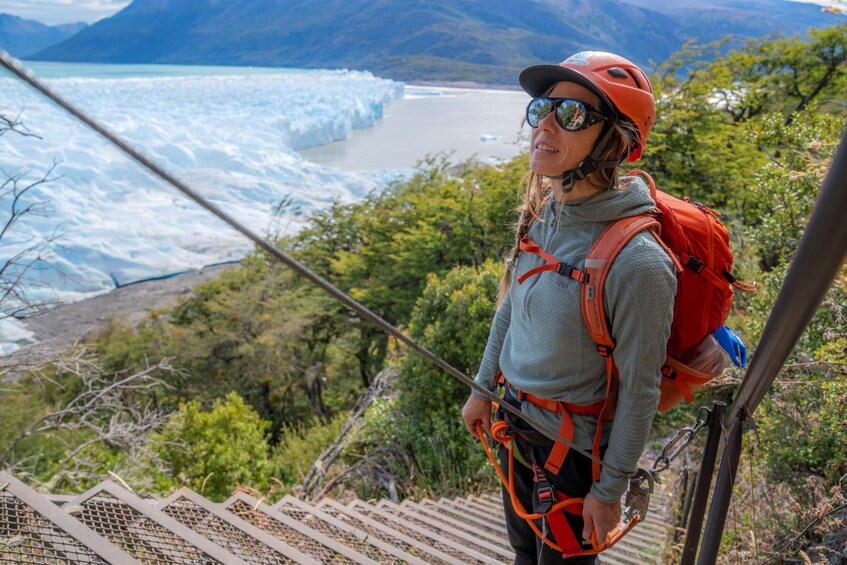 A person with a red backpack and helmet, standing on a wooden staircase overlooking a glacier and ocean. 3