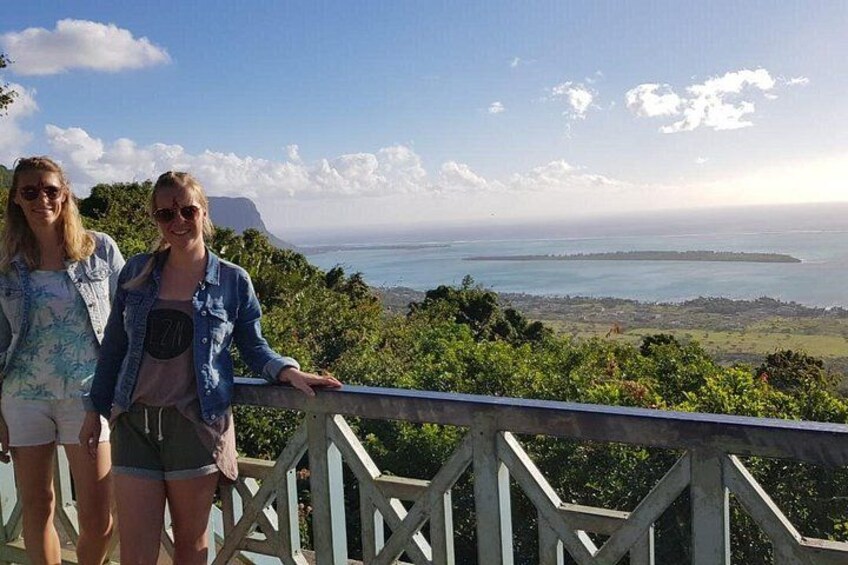Deux personnes se tiennent sur une terrasse en bois, d’où elles dominent un paysage côtier verdoyant et peuvent admirer la mer. 3