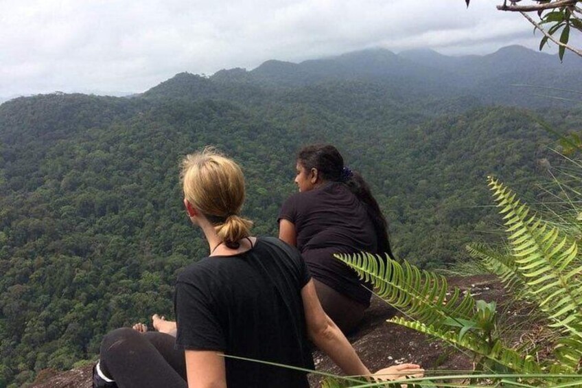 Two people sitting on a cliff overlooking a dense forested mountain range. 2