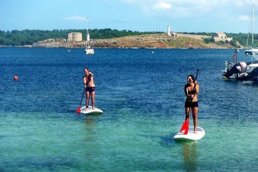 Two people paddleboarding on calm water with boats and a lighthouse in the background. 4