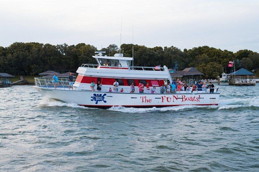 Ein weißes und rotes Boot mit dem Schriftzug 'The FUN Boats!', das sich auf dem Wasser bewegt, mit einer bewaldeten Uferlinie im Hintergrund. 3
