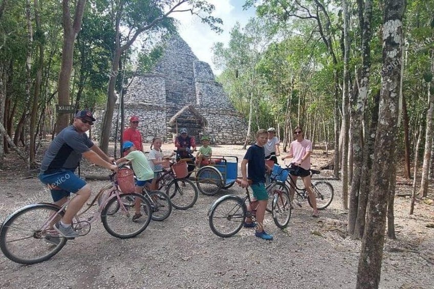 A group of people on bicycles in a forested area with a stone structure in the background. 1