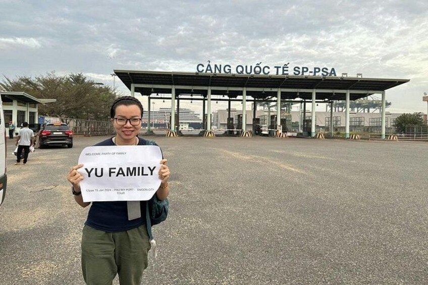 Eine Person hält ein Schild vor einer Mautstelle. Auf dem Schild stehen die Texte 'CÁNG QUỐC TẾ SP-PSA' und 'WELCOME PARTY OF FAMILY'. 2