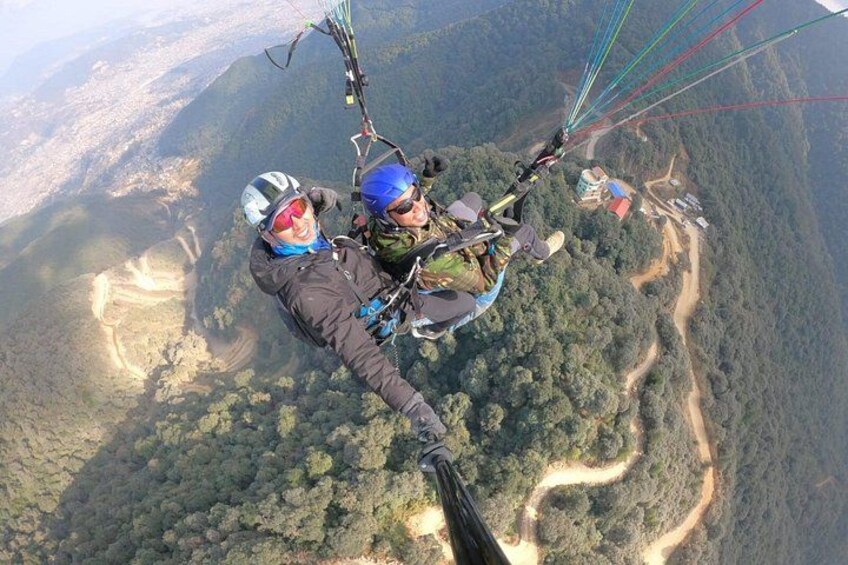 Two people paragliding over a mountainous landscape with dense forests and winding roads below. 5