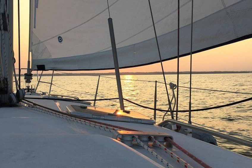 A sailboat with a white sail, a person standing on the deck, and the sun setting over the water. 1