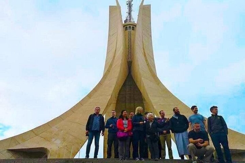 A group of people standing in front of a large, curved monument with a central spire. 2