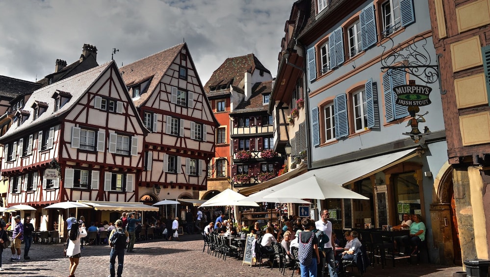A cobblestone square with traditional half-timbered buildings, outdoor cafes, and people dining. 2