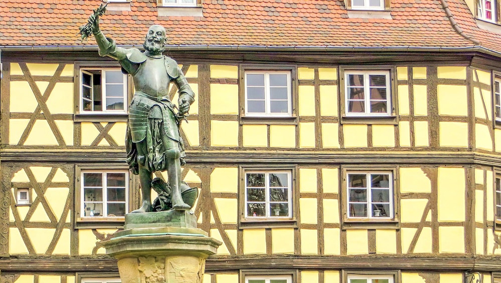 A statue of a historical figure in armor on a pedestal, set against a building with a yellow facade and timber framing. 1