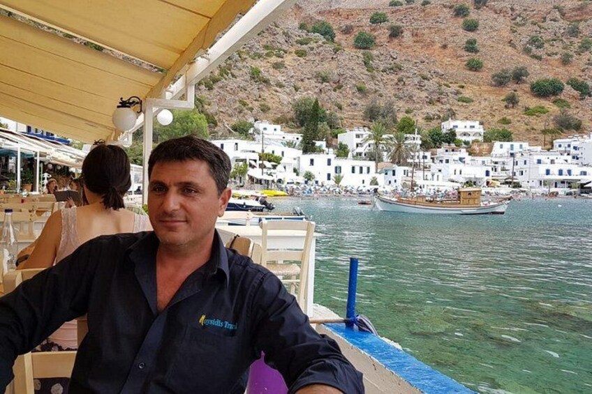 A man seated at a table on a boat, with a coastal town and hills in the background. 5