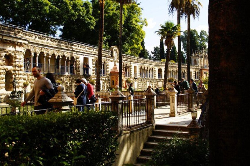 A historic courtyard with stone architecture, palm trees, and people walking. 5