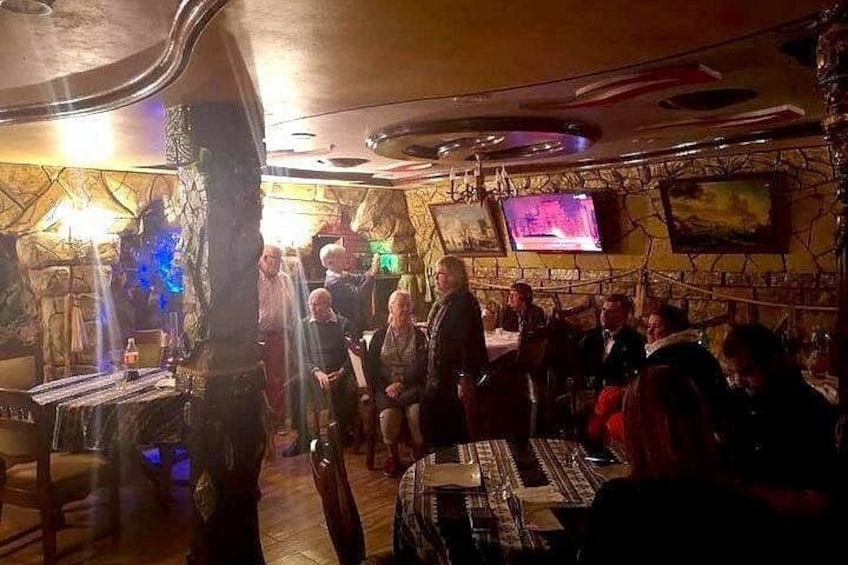 Un bar à la lumière tamisée, avec des clients assis à des tables, un téléviseur fixé au mur et un décor rustique caractérisé par des murs en pierre et des meubles en bois. 4