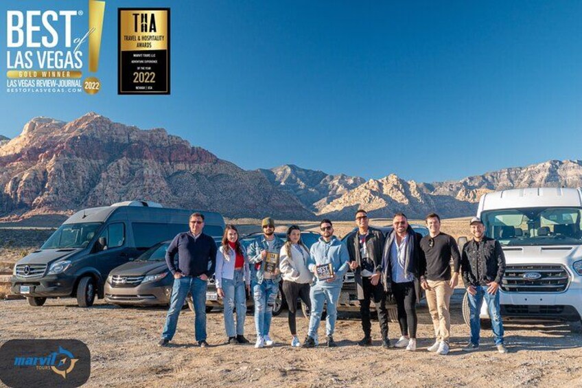 A group of people standing in front of a desert landscape with mountains in the background and two vehicles parked on the ground. 4