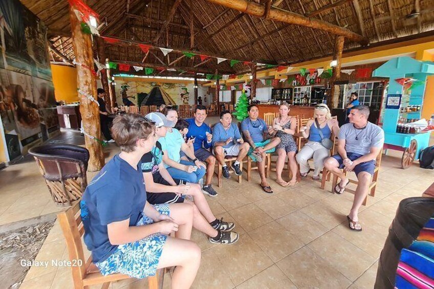 A group of people sitting in a circle on chairs in an outdoor setting with a thatched roof, wooden furniture, and a mural on the wall. 3