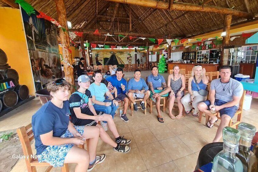 A group of people seated in a covered outdoor area with a thatched roof, wooden furniture, and a bar area in the background. 4