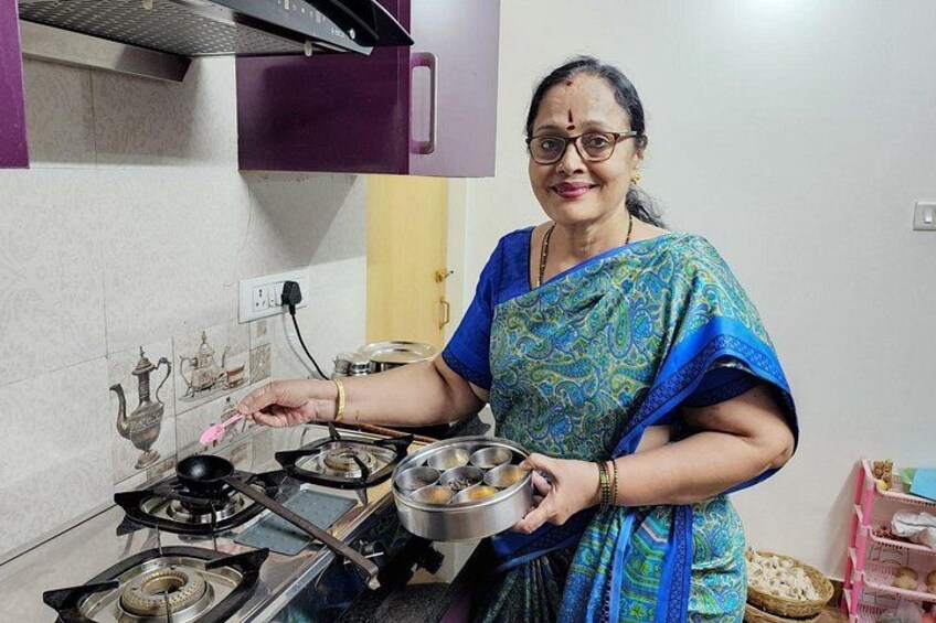 A person in a blue and green sari cooking in a kitchen with a gas stove, pots, and a teapot. 1