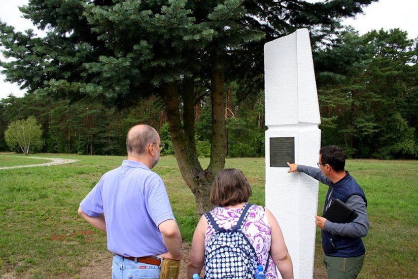 Tre personer der ser på et hvidt monument med en mindetavle i en park med træer. 1
