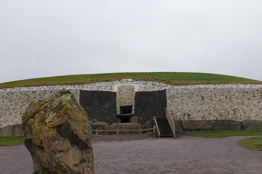 A stone structure with a staircase leading to a dark entrance, surrounded by a grassy hill and a large rock in the foreground. 1