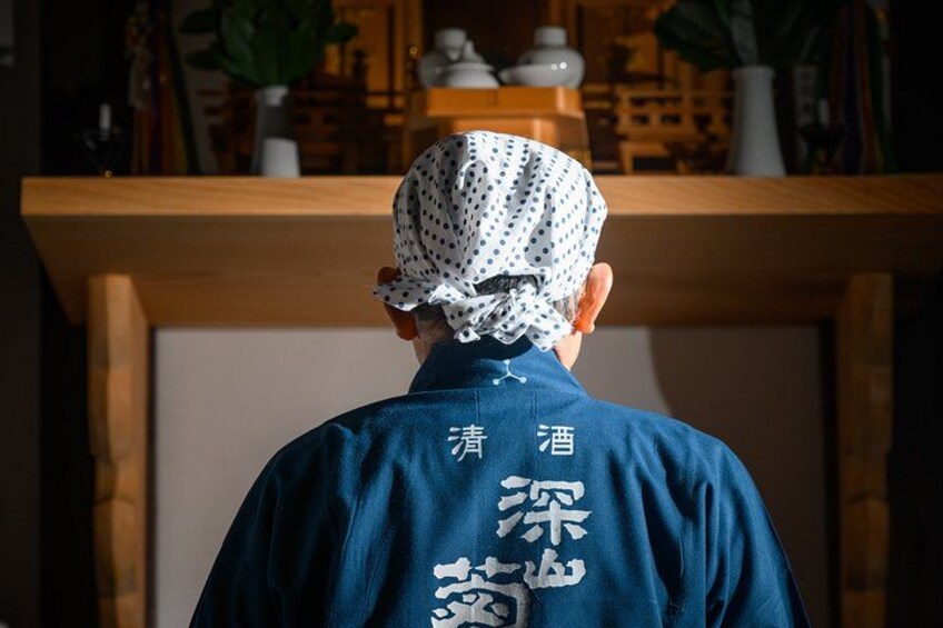 A person wearing a blue shirt with white text on the back, standing in front of a wooden counter with a plant and decorative items. 4