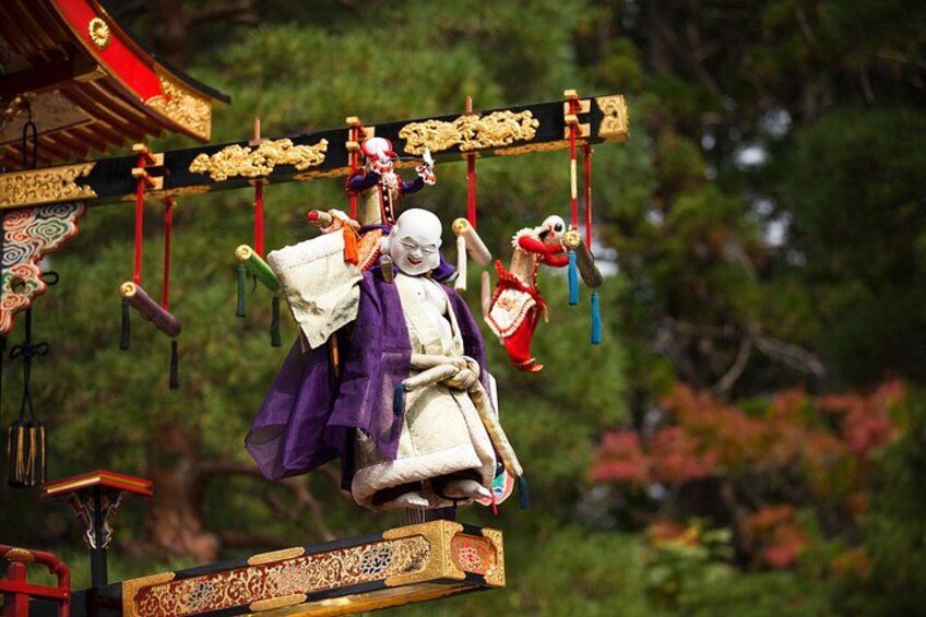 Ein traditionelles japanisches Puppentheater mit einer Puppe in einem violett-weißen Kostüm, vor einem Hintergrund aus Bäumen und einem roten Gebäude. 4