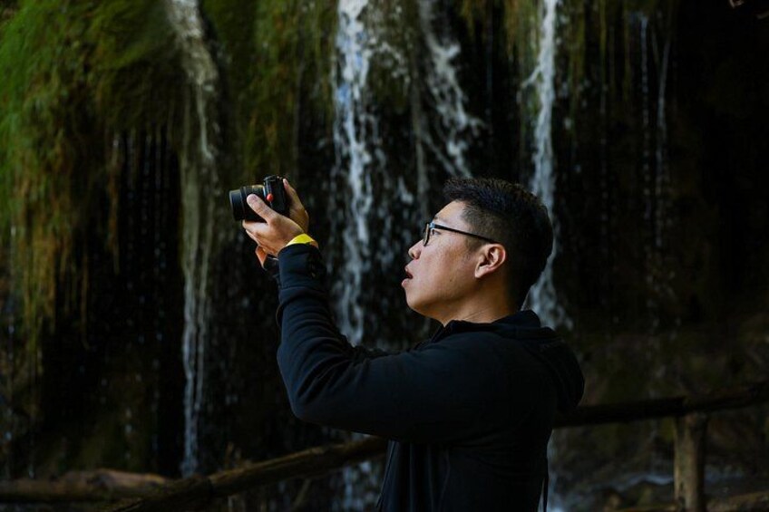A person taking a photo with a camera in a forested area with a waterfall in the background. 2