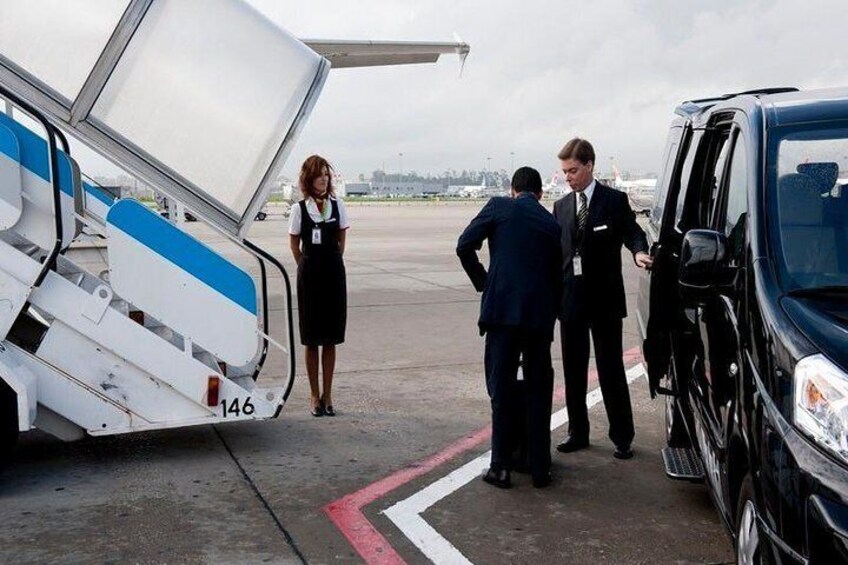 A person in a black suit standing next to a van, with an airplane partially open on the left. 3