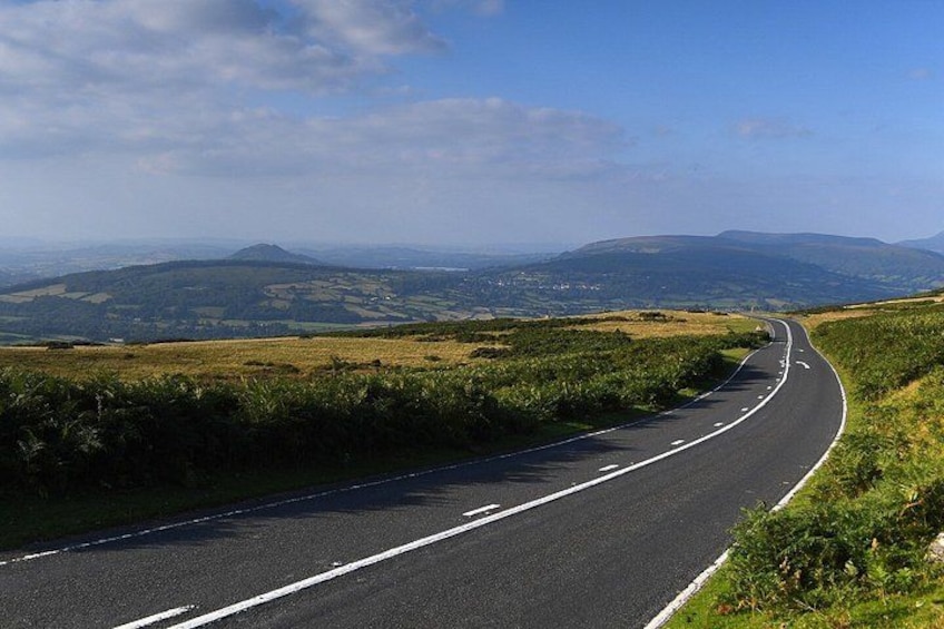 Una carretera sinuosa atraviesa un paisaje montañoso con campos verdes y montañas al lejano. 5