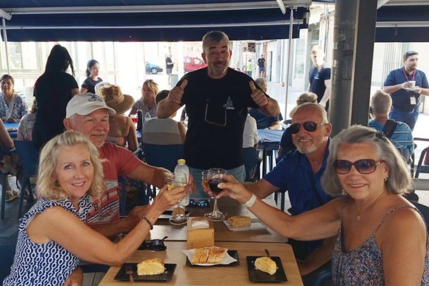 A group of people sitting at a table, toasting with drinks, in an outdoor setting with other diners in the background. 2