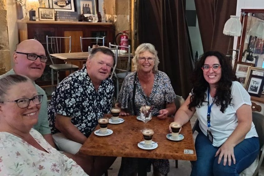 A group of people sitting around a wooden table with cups of coffee, in a room with framed pictures and curtains. 5