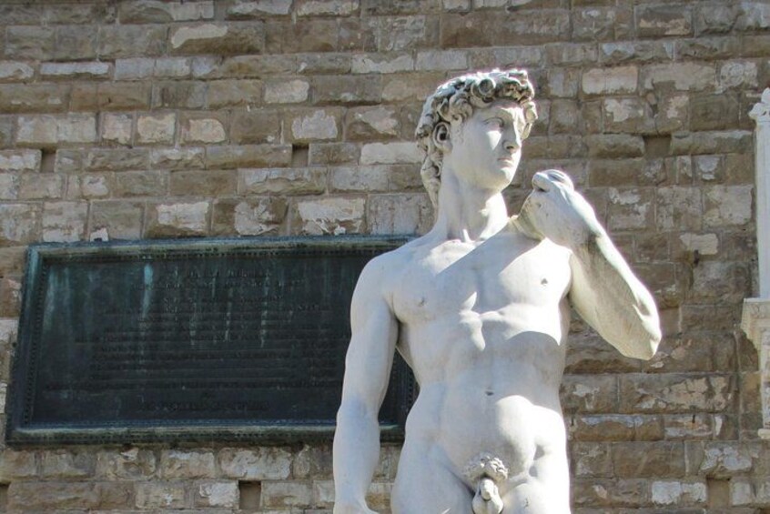 A classical statue of a male figure with a draped garment, standing against a stone wall with a metal plaque. 2