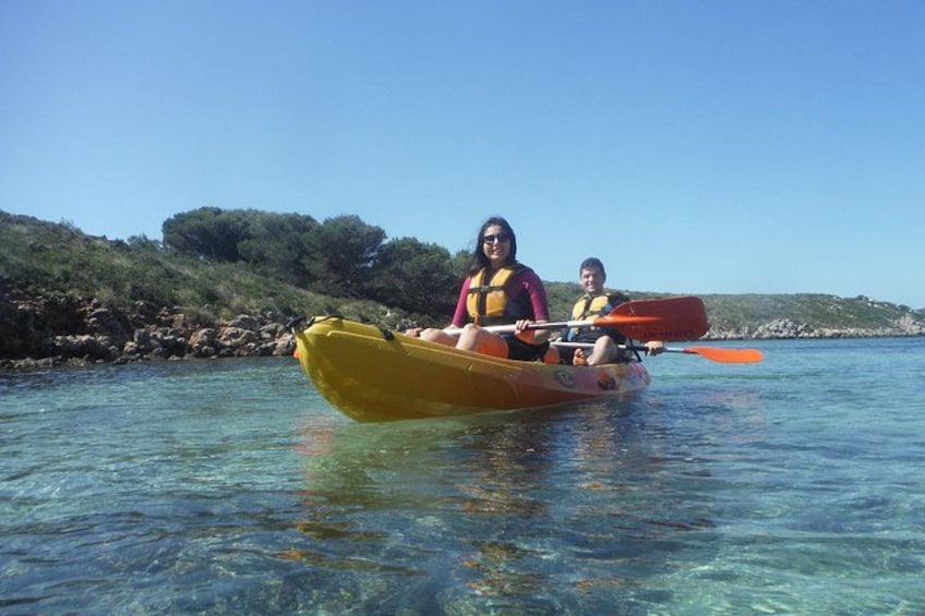Two people kayaking on clear water with a rocky shoreline and greenery in the background. 1