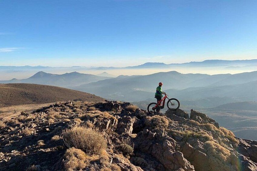 En person cykler på en mountainbike over ujævnt terræn med udsigt til fjerne bjerge og dale. 2