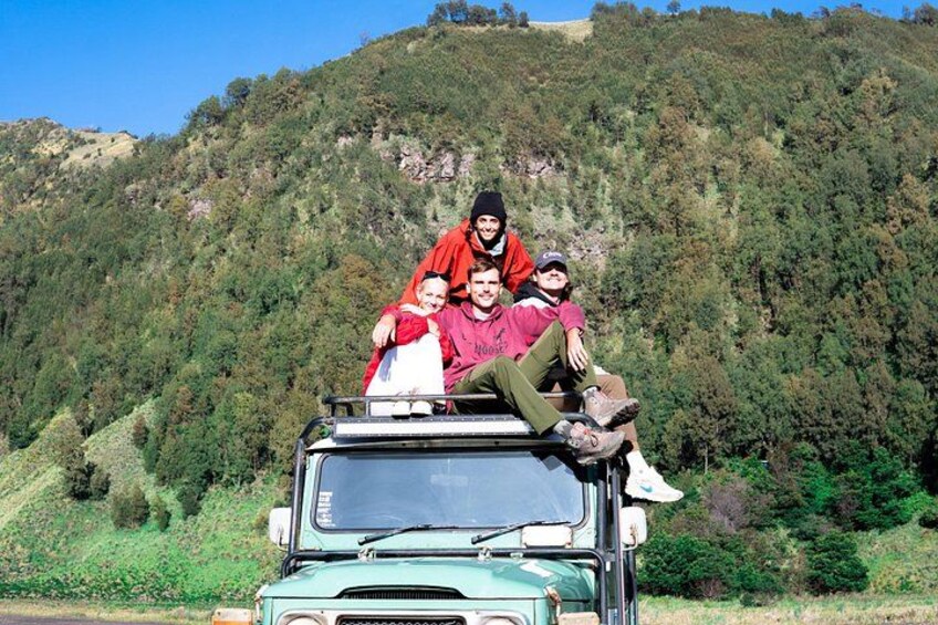 A group of people sitting on the roof of a green vehicle, with a forested hillside in the background. 3