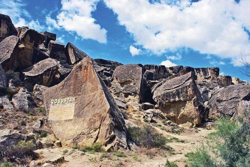 Een rotsachtig landschap met een grote rots waarop 'QOBUSTAN' staat, en een helderblauwe lucht. 1
