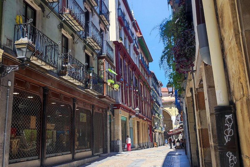 A narrow street with multi-story buildings, balconies, and a pedestrian. 3