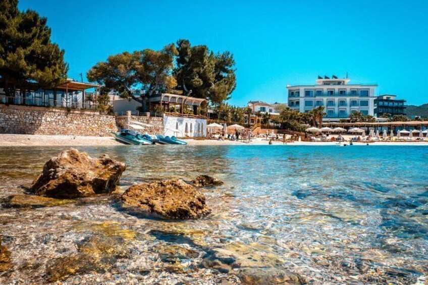 A beach with clear water, rocks, and buildings in the background. 5