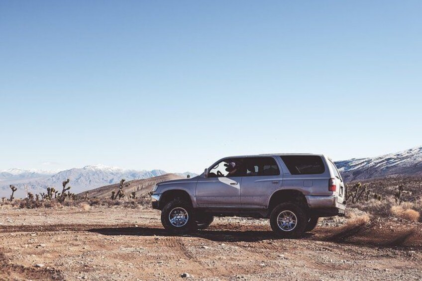 A silver SUV parked on a dirt road with a desert landscape and mountains in the background. 1