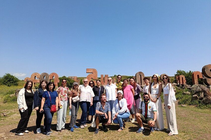 A group of people posing in front of large, rusted letters spelling out a name on a grassy field. 3