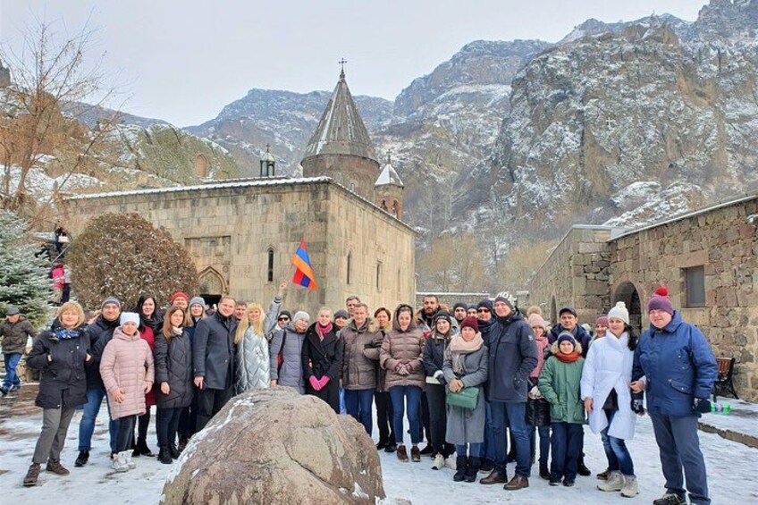 A group of people standing in front of a church with a flag, snow-covered mountains in the background. 2