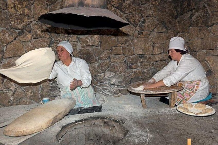 Two people in a stone-walled kitchen, one kneading dough and the other preparing a flatbread. 3