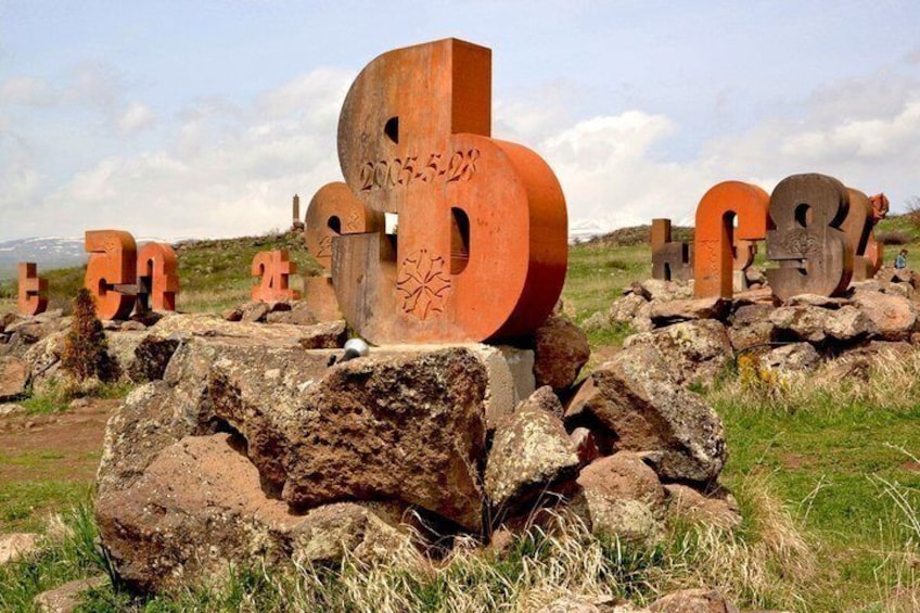 Large, rusted metal letters with numbers on rocks in a grassy field. 2