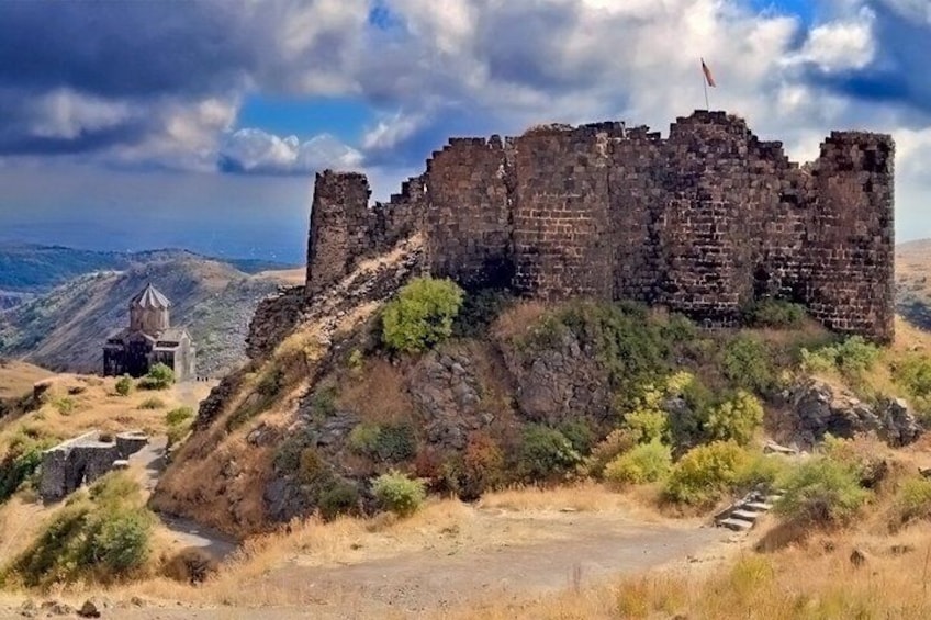 A historic stone fortress with ruins, a small chapel, and a flag on top, surrounded by hills and dry vegetation. 5