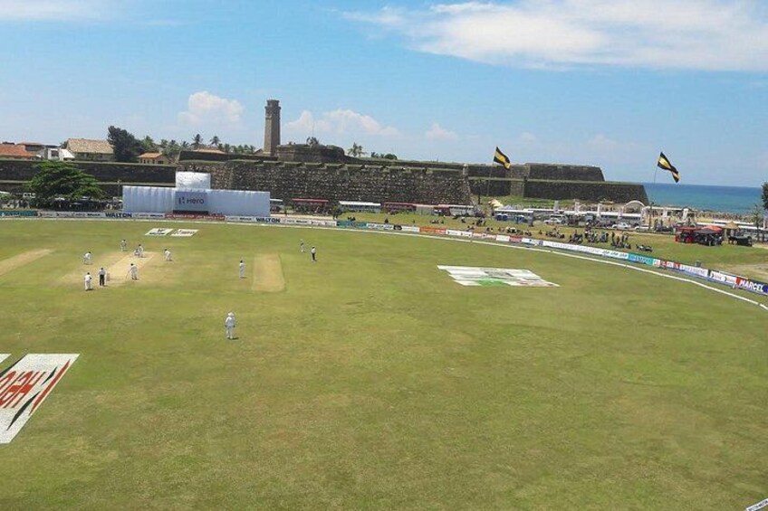 A cricket field with players, a large banner, and a coastal backdrop with a fort. 4