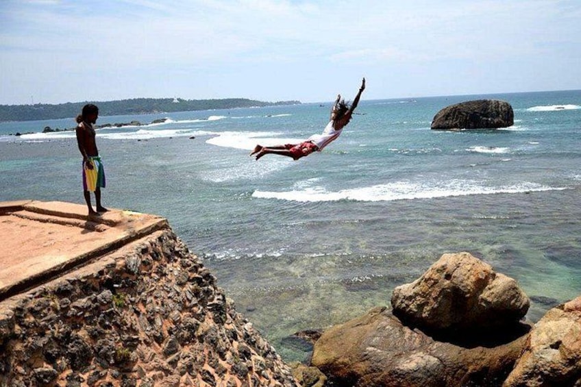 A person jumping off a cliff into the ocean, another person watching, and rocky terrain with clear water. 5