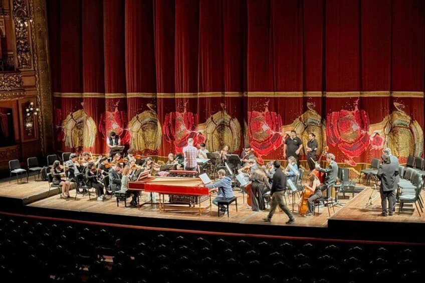 A theater stage with orchestral musicians, a conductor, and a conductor's podium. 4