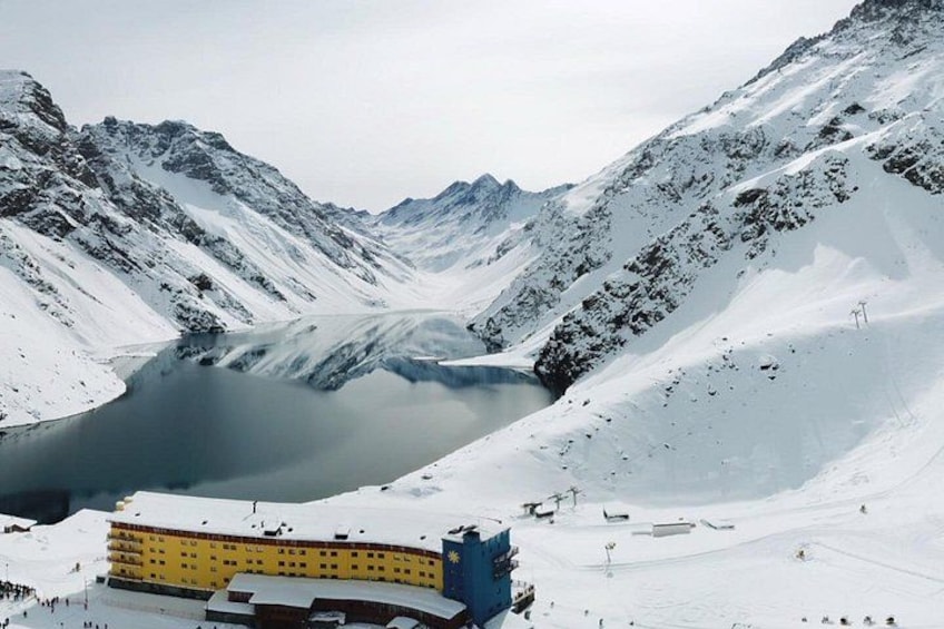Un paisaje montañoso nevado con un lago, un edificio amarillo y pistas de esquí. 1