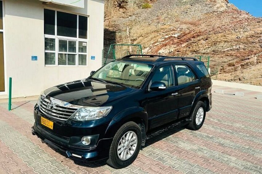 Ein schwarzer SUV parkt auf einem befestigten Untergrund, im Hintergrund ein Gebäude und ein felsiger Hügel. 3