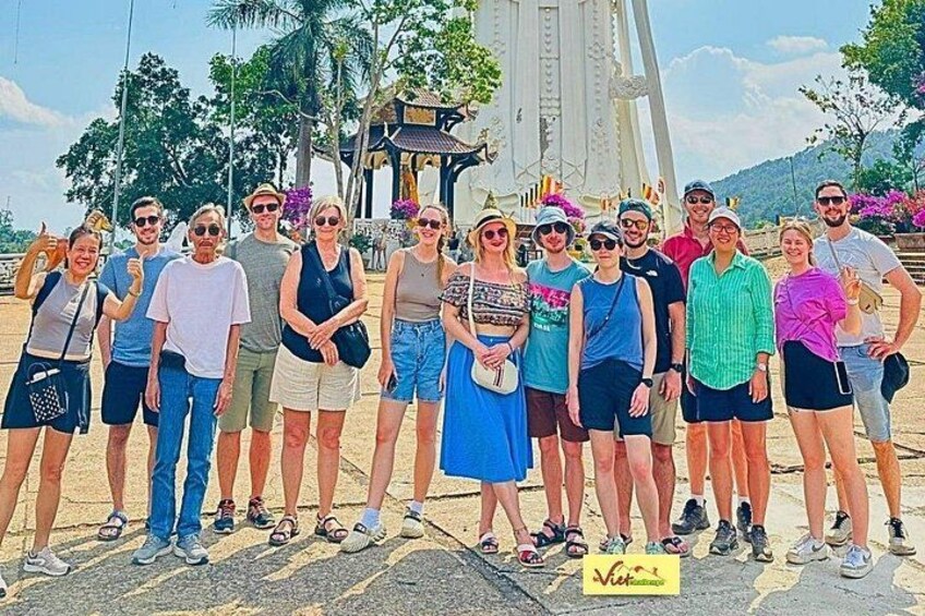 Un groupe de personnes posant devant une grande structure blanche surmontée d’un toit en forme de pagode, avec des palmiers et un ciel dégagé. 2