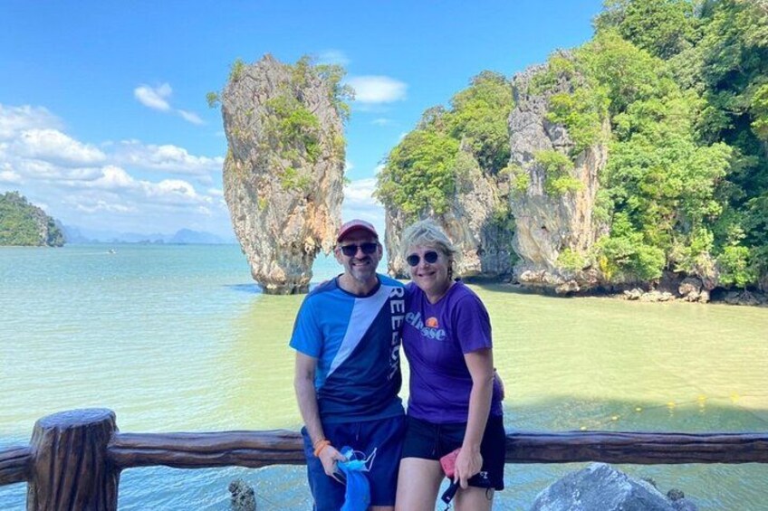 Two people standing by a wooden railing with a clear view of a large rock formation in the water and a forested island in the background. 5